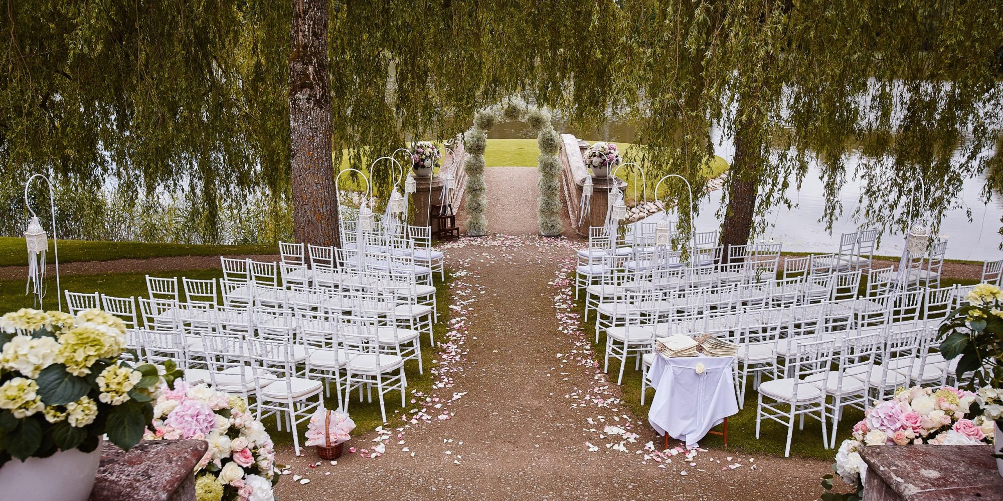 Place for wedding ceremony with wedding arch decorated with flowers and white chairs on each side of archway outdoors. Preparation for wedding ceremony outdoors near lake.